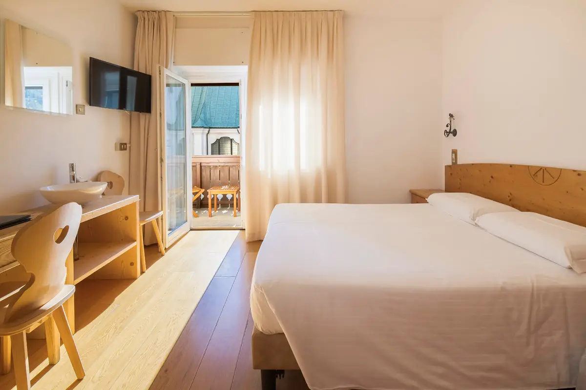 Camera Family con balcone soleggiato e zona notte luminosa, arredata in stile alpino a Cortina d'Ampezzo. | Family room with sunny balcony and bright sleeping area, furnished in alpine style in Cortina d'Ampezzo.