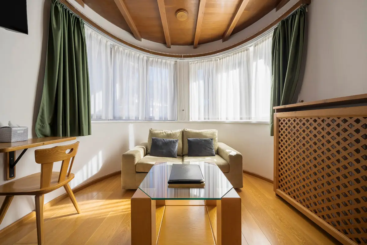Zona giorno con finestra curva nella camera Superior Tripla – Hotel Montana Cortina. | Curved-window sitting area in the Superior Triple Room – Hotel Montana Cortina.